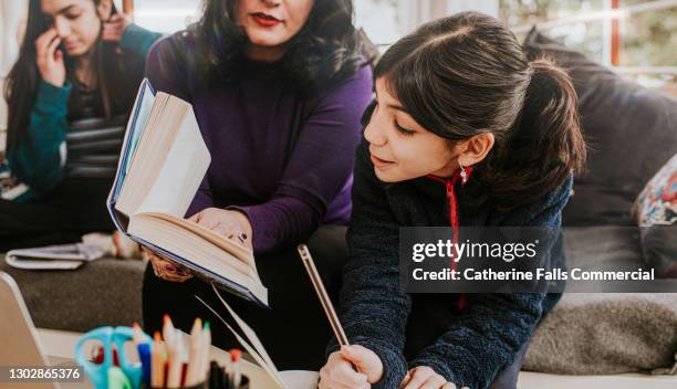 a mother helps her younger daughter with her homework while older teenager reads in the background - english language fotografías e imágenes de stock