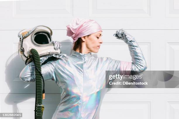 sick woman with breast cancer dressed as an astronaut with a helmet in her hand, wearing a pink head scarf and armband, making a symbol of strength - best bosom stock-fotos und bilder