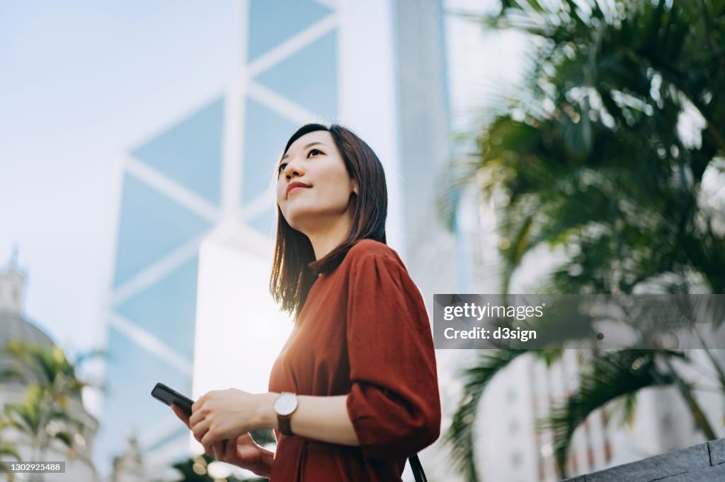 Confident and determined young Asian businesswoman looking away hopefully, using smartphone on the go against contemporary corporate skyscrapers in central business district in the city. Female leadership and determined to success