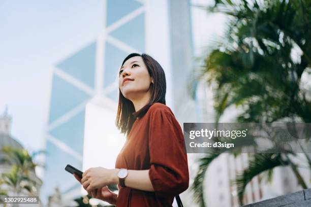 confident and determined young asian businesswoman looking away hopefully, using smartphone on the go against contemporary corporate skyscrapers in central business district in the city. female leadership and determined to success - financial aspirations stock pictures, royalty-free photos & images