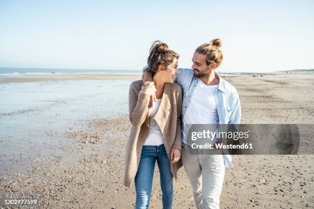 young smiling couple looking at each other while walking on beach during weekend - young couple beach stock pictures, royalty-free photos & images