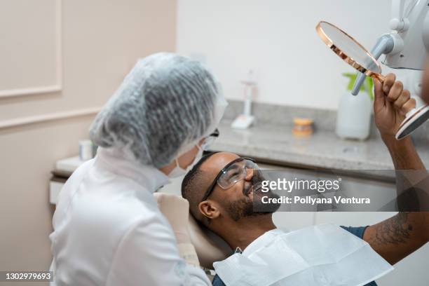 hombre en silla de dentista mirando los dientes en el espejo - odontología cosmética fotografías e imágenes de stock