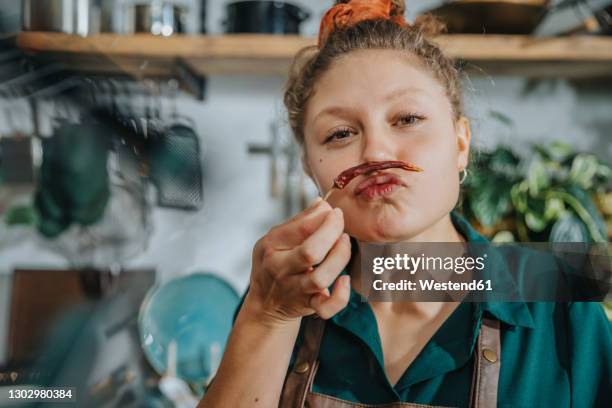 playful chef making mustache with dry chili pepper while standing in kitchen - chili woman fotografías e imágenes de stock