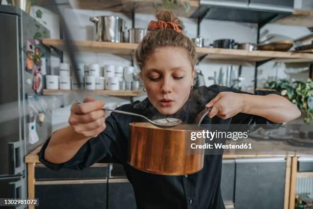 young female chef tasting broth soup while standing in kitchen - pots and pans stock-fotos und bilder
