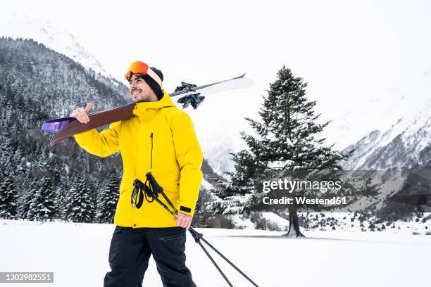 young man with ski looking away while walking on snow covered valley - skijacke stock-fotos und bilder