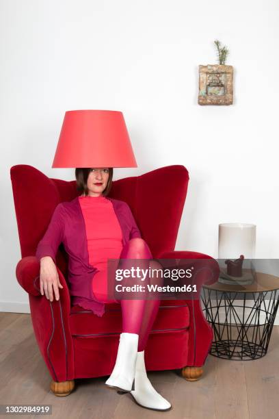 mature woman with lamp shade on head sitting on armchair against white wall - lamp shade fotografías e imágenes de stock