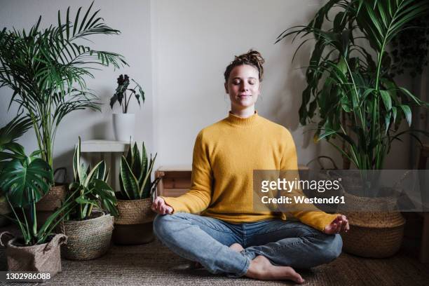 young woman meditating while sitting on floor by plant at home - tranquilidad fotografías e imágenes de stock