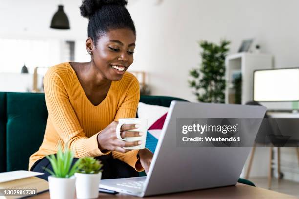 happy afro woman holding cup while using laptop at home - couchtisch stock-fotos und bilder