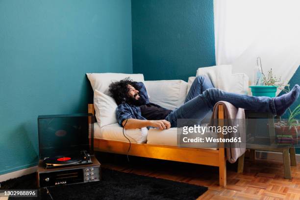 bearded man listening music through turntable while lying on sofa at home - man living room fotografías e imágenes de stock