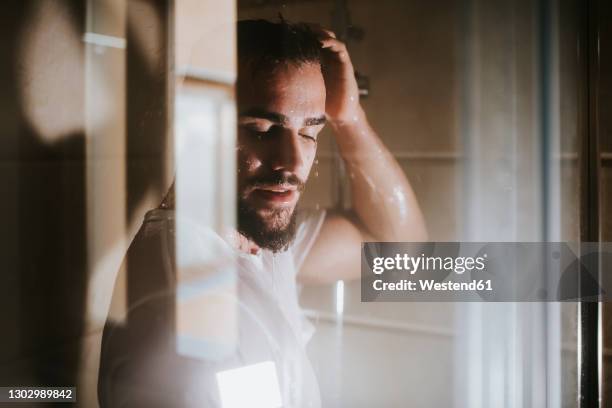 young man with eyes closed taking shower seen through glass in bathroom - bath shower stock pictures, royalty-free photos & images