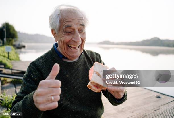 happy man showing thumbs up while eating sandwich sitting on pier - grundnahrungsmittel stock-fotos und bilder