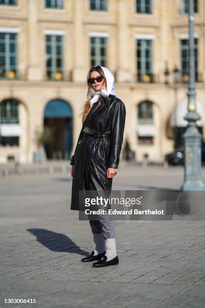 Natalia Verza wears a white scarf over the head, sunglasses, a black leather long trench coat, a white turtleneck top, gray jeans, gray wool socks...