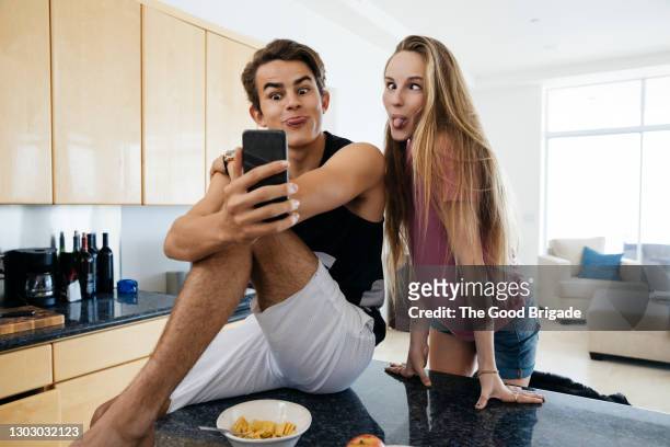 young man and sister making face while photographing selfie in kitchen - sibling stock pictures, royalty-free photos & images