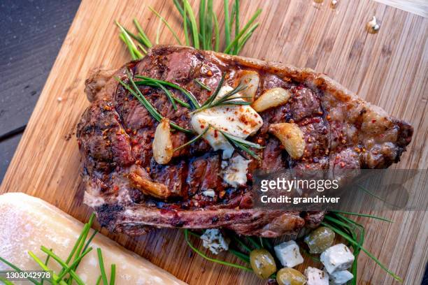 ribeye steak just off a charcoal grill resting on a wood cutting board, seasoned with rosemary, garlic cloves, butter, cheese, olives and paprika - rib eye steak stock pictures, royalty-free photos & images