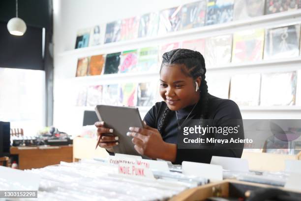 an african american millennial woman with french braids in her hair, reads from a digital tablet and wears ear pods in a record store while wearing a black turtleneck and jeans. - chicago lifestyle stock pictures, royalty-free photos & images