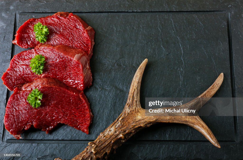 Raw steak meat from roe deer on the bridlic chopping board. Roe deer antler as a decoration. Copy space for text.