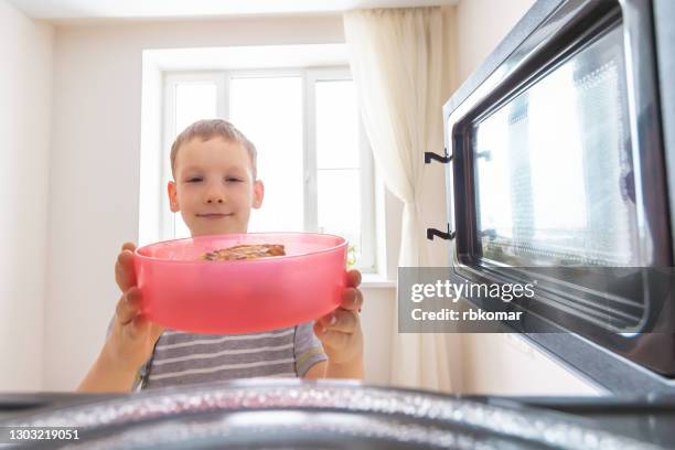 a happy child puts a bright plastic container of food in the microwave to reheat with the door open. inside view of a boy alone in the kitchen - hungrybox stock pictures, royalty-free photos & images