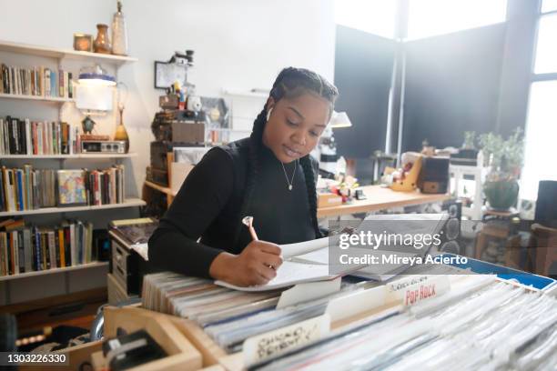 an african american millennial woman with french braids in her hair, uses her textbook to work on her homework in a small record store while wearing a earbuds, a black turtleneck and jeans. - black turtleneck stock pictures, royalty-free photos & images
