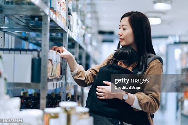 young asian mother doing grocery shopping in an organic grocery store while carrying a sleeping baby. healthy eating and shopping lifestyle - mãe dona de casa imagens e fotografias de stock