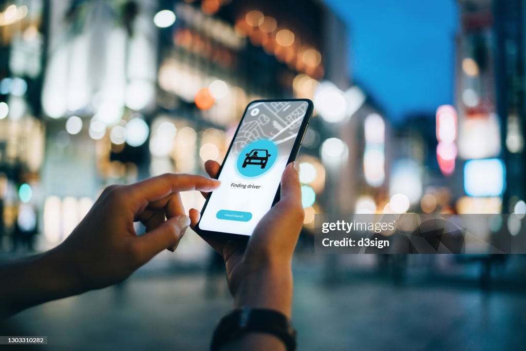 Close up of young woman using mobile app device on smartphone to arrange a taxi ride in downtown city street, with illuminated city traffic scene as background
