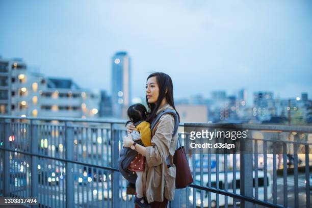 young asian working mother commuting in the city on urban bridge after work while carrying little daughter, against illuminated city skyline in the evening. domestic life of a working mother. concept of work-life balance - fukuoka city photos et images de collection
