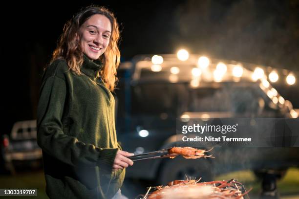 young woman flipping shrimp for barbecue grill party over open campfire. camping car lifestyle, sustainable lifestyle - night picnic stock pictures, royalty-free photos & images