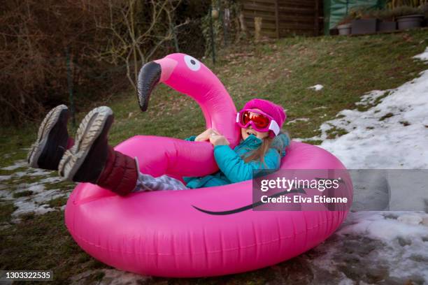 child using an inflatable pink flamingo to slide down a slope of receding winter snow in a back yard - child slide stockfoto's en -beelden