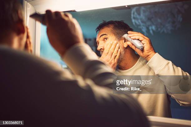 friendly man cuts his own hair with a machine in front of the mirror during coronavirus lockdown. - beard trimming stock pictures, royalty-free photos & images