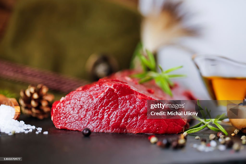 Raw steak meat from deer on the bridlic chopping board with ingredients as a sea salt, pepper , rosemarine, oil and hunting hat as a decoration.