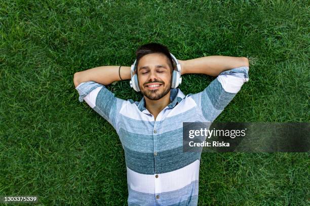 junger mann mit kopfhörern auf dem rasen im park liegen und entspannen - im gras liegen stock-fotos und bilder