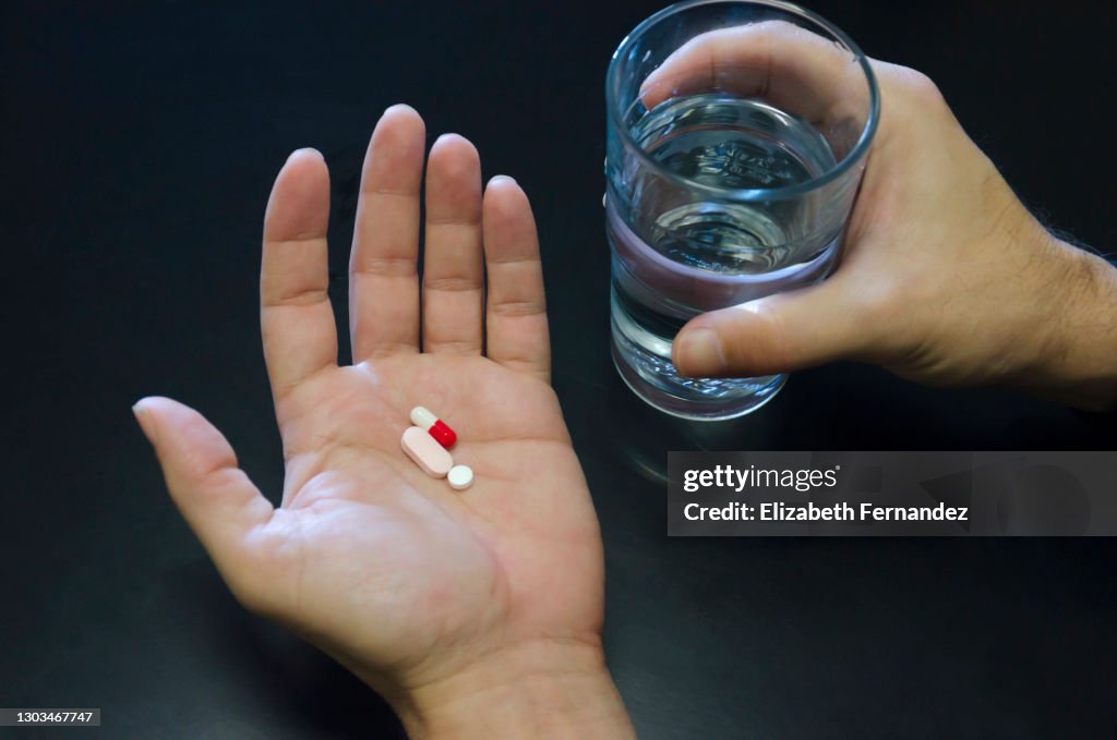 Hands holding three pills and a glass of water against black background, copy space