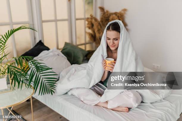 morning, comfort and people concept - young woman with cup of coffee sitting in bed under blanket at home bedroom - wrapped in a blanket stock pictures, royalty-free photos & images