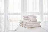 Stack of fresh white bath towels on bed sheet in modern hotel bedroom interior with window on background, copy space