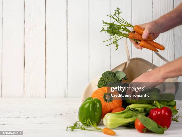 many fresh market vegetables in a natural cotton reusable shopping bag on an old weathered white wood panel table against a white paneled wood wall, with a person removing some vegetables from the bag. - arm made of vegetables stock pictures, royalty-free photos & images