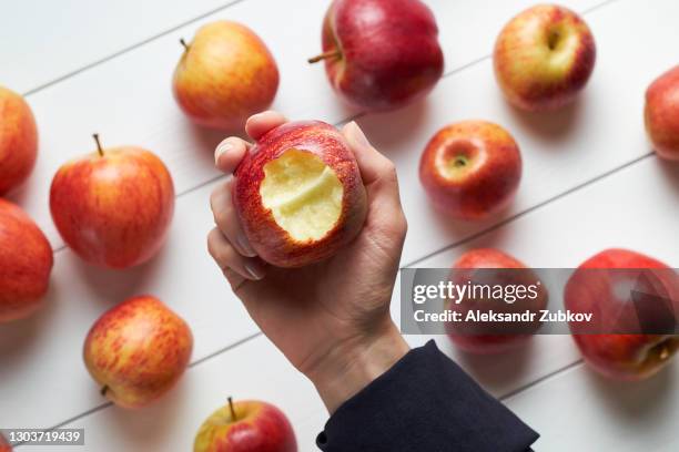 red ripe apples on a white wooden background. a woman holds a bitten apple in her hand. vegetarian, vegan, raw food. organic farm products. - eaten stock pictures, royalty-free photos & images