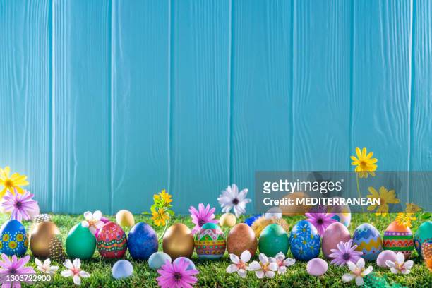 easter eggs on turf grass and blue wooden wall with spring flowers - easter stock pictures, royalty-free photos & images