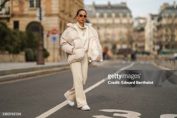 Camille Coyere wears sunglasses, a white oversized winter puffer long coat, a white pullover with a front zipper, white flared pants, white Nike...