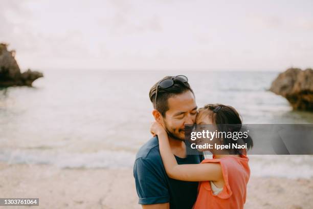 happy father and young daughter cuddling on beach at sunset, japan - asian father and daughter stock pictures, royalty-free photos & images