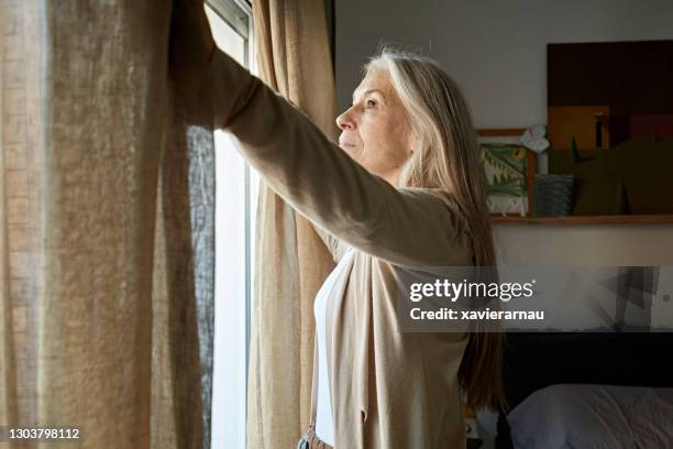 mid 60s caucasian woman opening curtain in bedroom - opening the curtains stock pictures, royalty-free photos & images