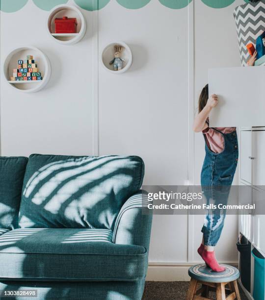 a little girl stands on her tip-toes on a little stool to open a cabinet that is too high to reach otherwise - skimpy girls stock pictures, royalty-free photos & images