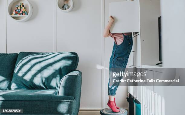 a little girl stands on her tip-toes on a little stool to open a cabinet that is too high to reach otherwise - neugierde stock-fotos und bilder