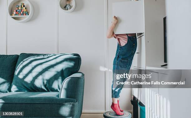 a little girl stands on her tip-toes on a little stool to open a cabinet that is too high to reach otherwise - cabinet bildbanksfoton och bilder