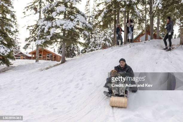 Father and daughter ride wooden sled down snowy hill while spectators watch