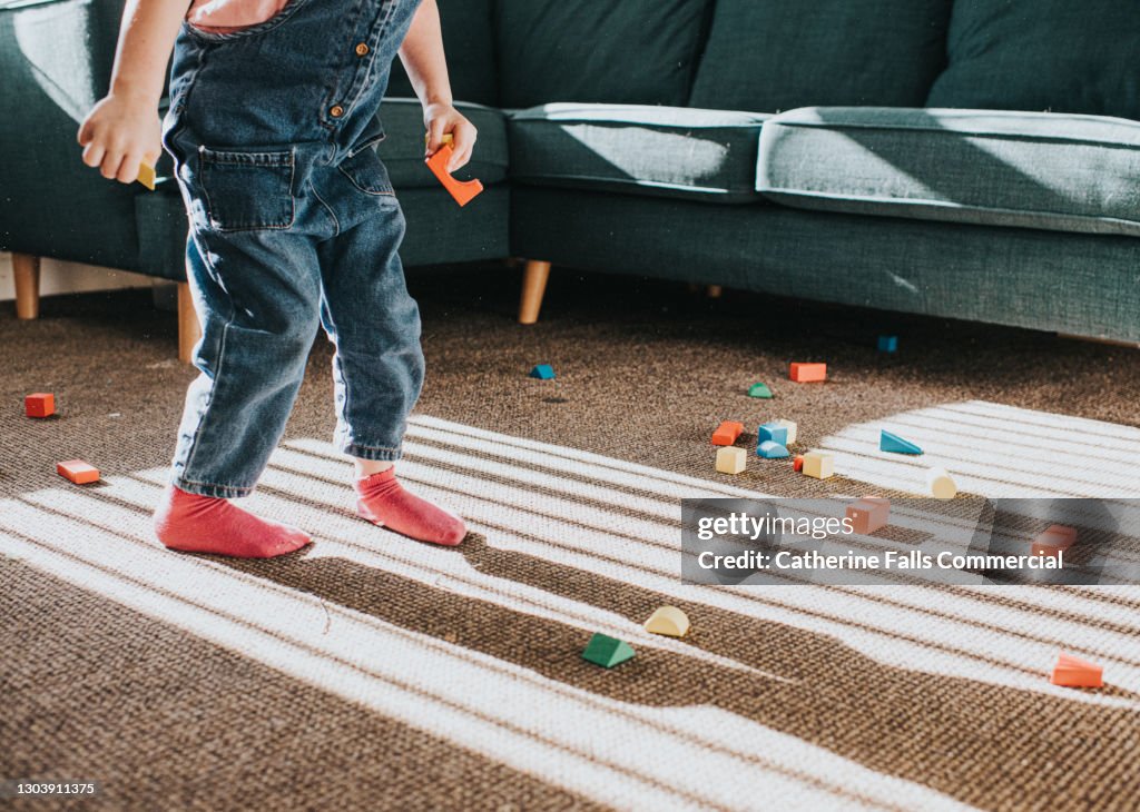 Child stands on carpet in front of a corner sofa. Wooden building blocks are scattered over the floor.