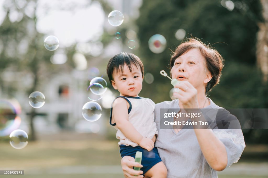 Asian chinese grandmother playing bubble together with her grandson in public park during weekend evening backlit