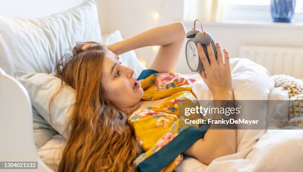 surprised blond young woman in bed late in the morning, checking the alarm clock and having overslept, being late for work - overslept stock pictures, royalty-free photos & images