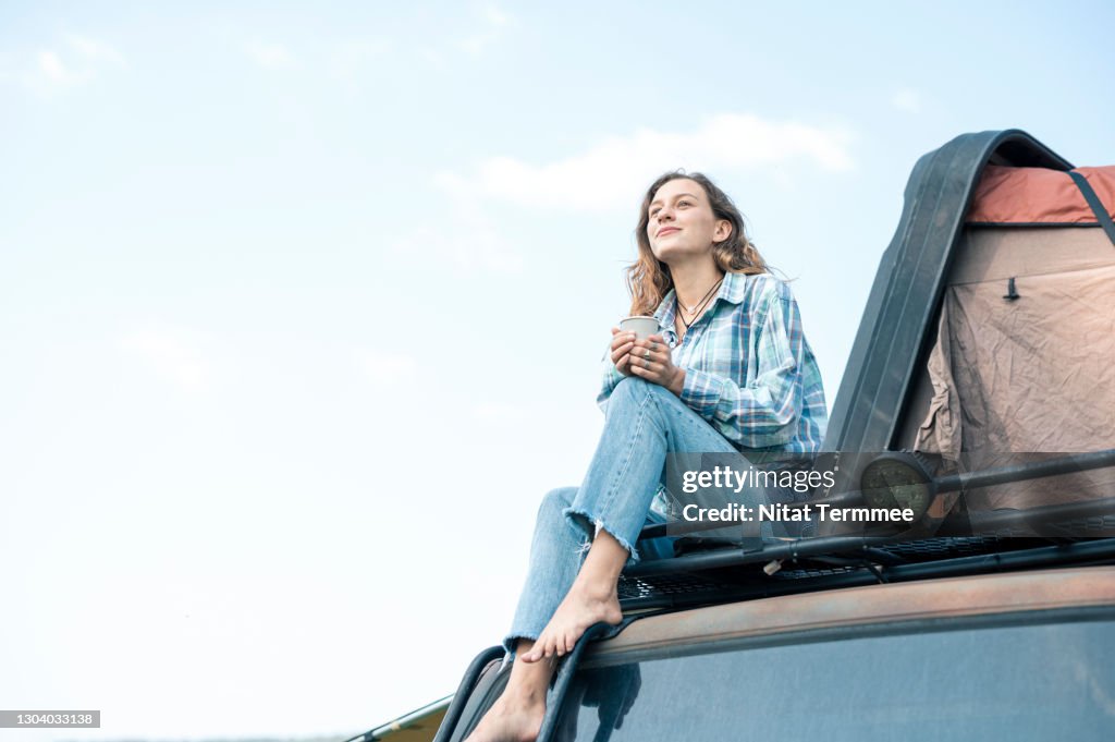 Portrait of Happy woman sitting on car roof of van while holding reuse coffee cup and enjoy the view. Camping car, Sustainable Lifestyle.