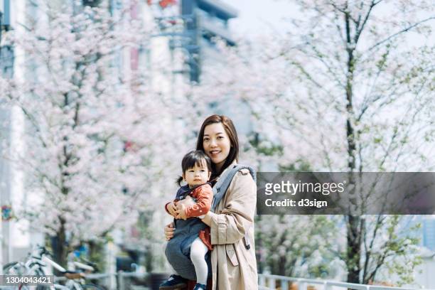portrait of smiling young asian mother and adorable little daughter under japanese cherry blossoms on a beautiful spring day. enjoying intimate family bonding time together. family lifestyle and nature - japanese mom stock-fotos und bilder