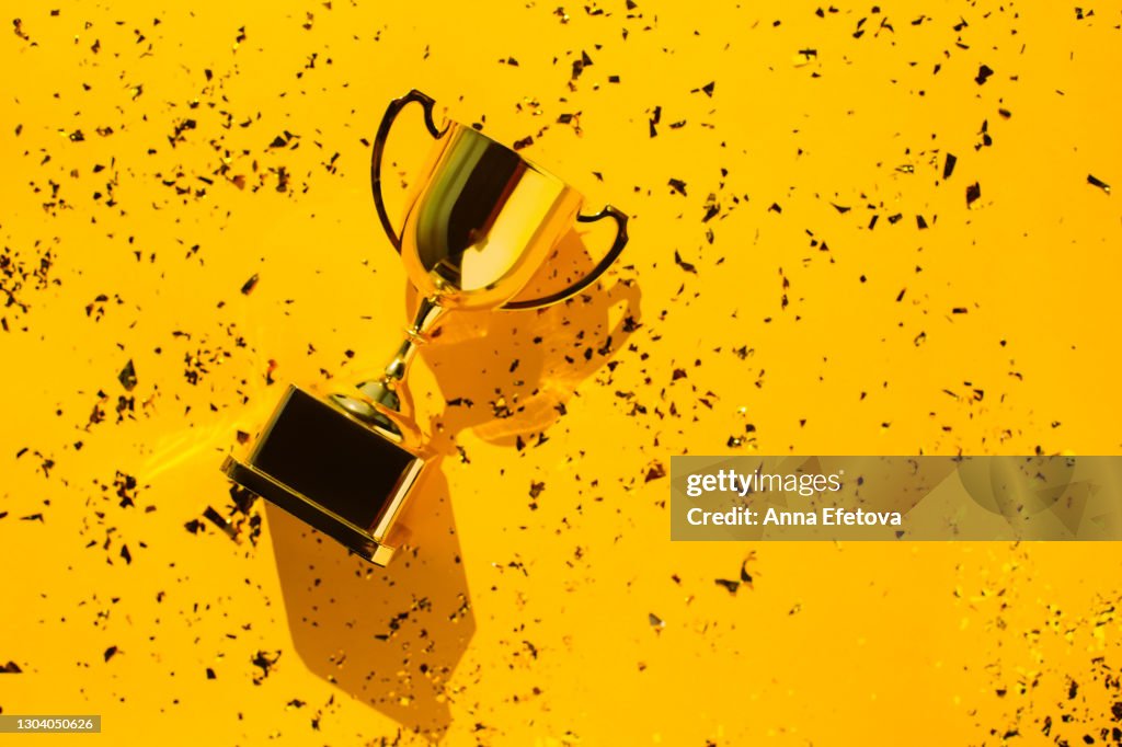 Top view of metallic golden goblet on bright yellow background with sequin. Goal achievement concept. Trendy colors of the year