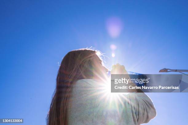blonde woman drinking water outside while being illuminated by a ray of sunlight. - calentador fotografías e imágenes de stock